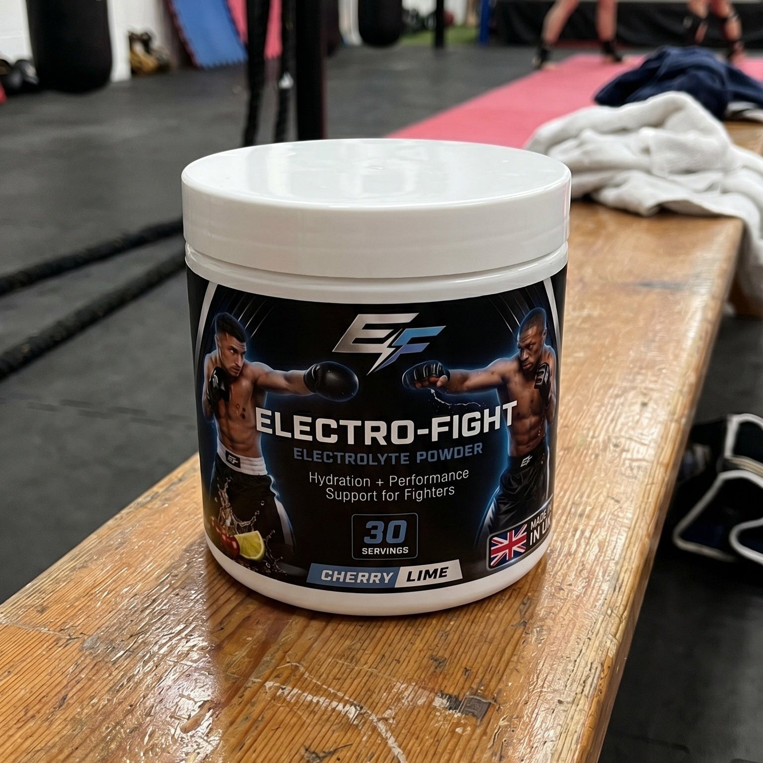 A tub of Electro-Fight Electrolyte Powder sits on a wooden gym bench with a boxing gym in the background.