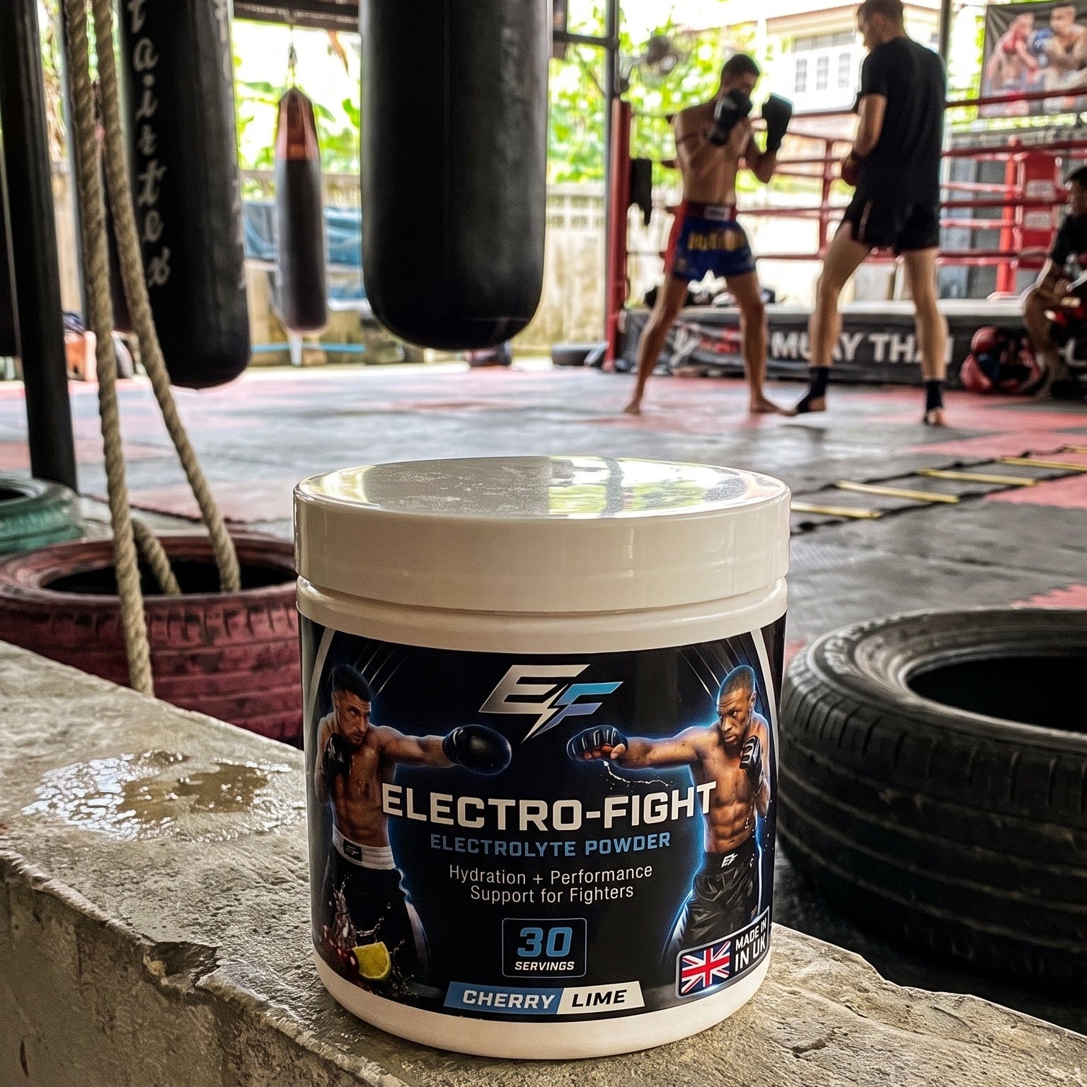 A tub of Electro-Fight Electrolyte Powder supplement placed on a concrete ledge in a boxing gym, with punching bags, a ring, and people training in the background.